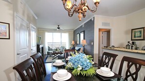 A dining area with a glass table, chairs, and a chandelier. There is a kitchen area with a counter and appliances visible in the background. - The Palm Tree Penthouse (Stuart)