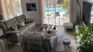 Een woonkamer met een bank, fauteuil, salontafel en voetenbankje. Er is een groot raam met gordijnen en uitzicht op een zwembad.