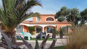 Een huis met een pannendak, een terras met stoelen en een palmboom op de voorgrond.