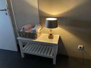 A small white bedside table with a lamp and a wicker basket containing rolled towels.