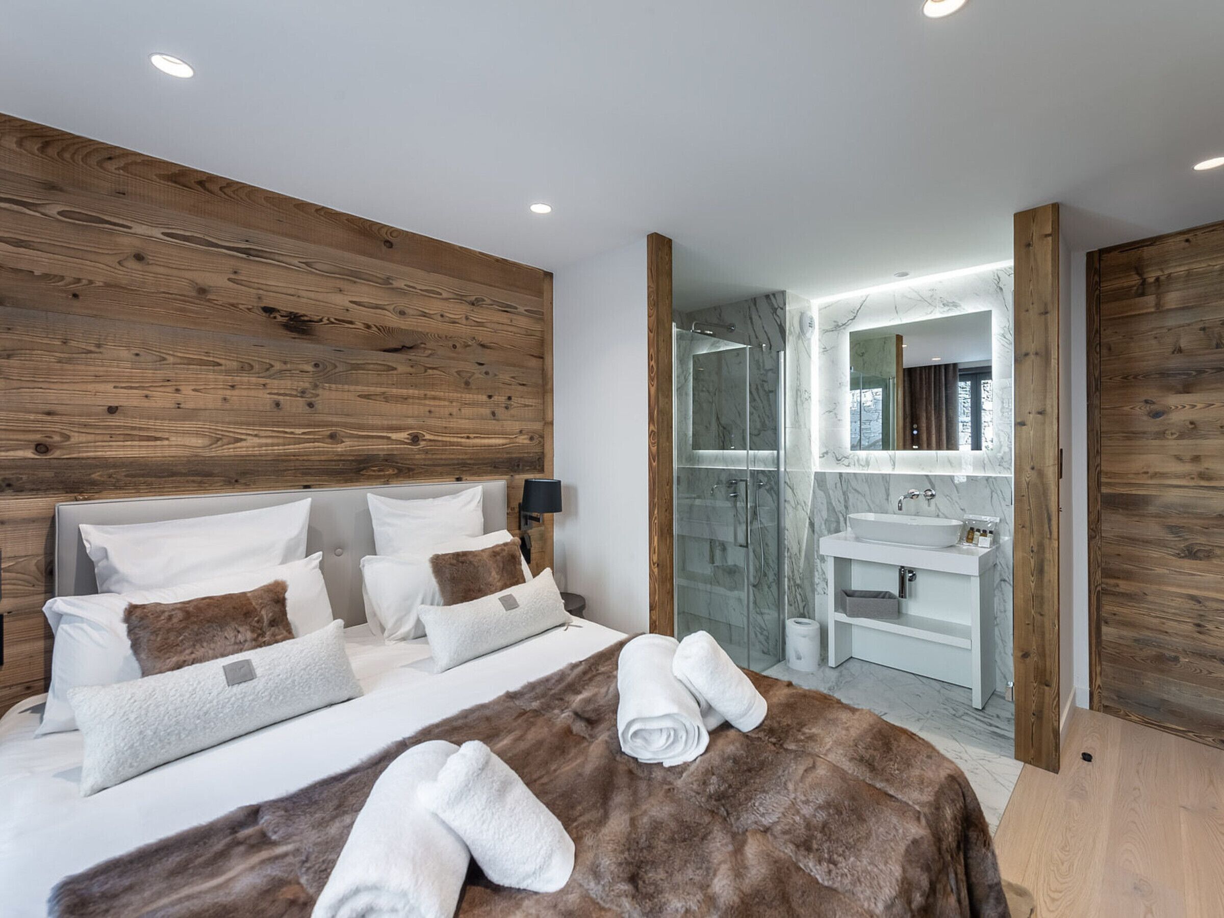 Quarto moderno com cabeceira de madeira, box de vidro para o chuveiro e toucador de banheiro em mármore.