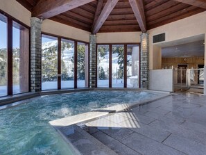 Een wellnessruimte met een bubbelbad, stenen pilaren, grote ramen en een uitzicht op besneeuwde bergen.