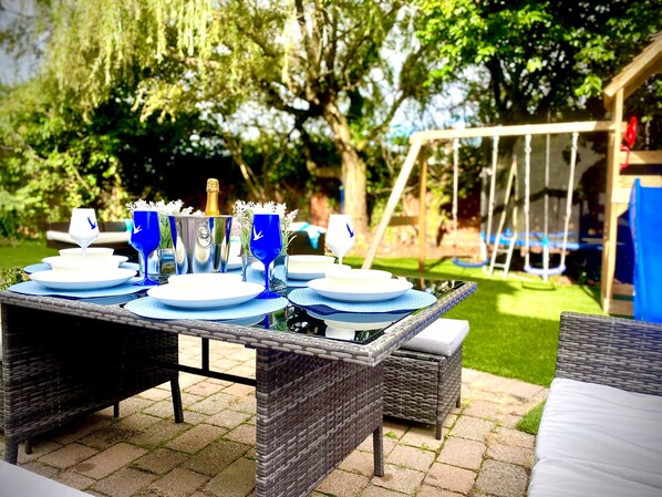 An outdoor dining area with a table set for four, surrounded by wicker furniture and a swing set in the background. - Anchor Hot tub Suite in Lytham St Annes (Lytham St. Anne's)