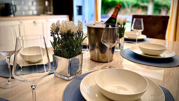 Une table à manger dressée avec de l’assiette blanche, des bols et des verres à vin, un vase  à fleurs et une bouteille de champagne dans un seau en métal.