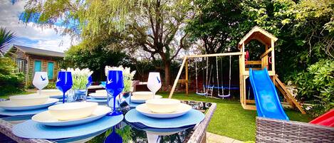 A backyard with a dining table set for six, surrounded by a small pond and a wooden play structure with a slide.