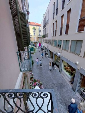 A narrow pedestrian street with buildings on either side, a shop on the right, and a balcony with a wrought iron railing in the foreground. - LA PRU House - Centro Storico - Mini Loft (Novara)