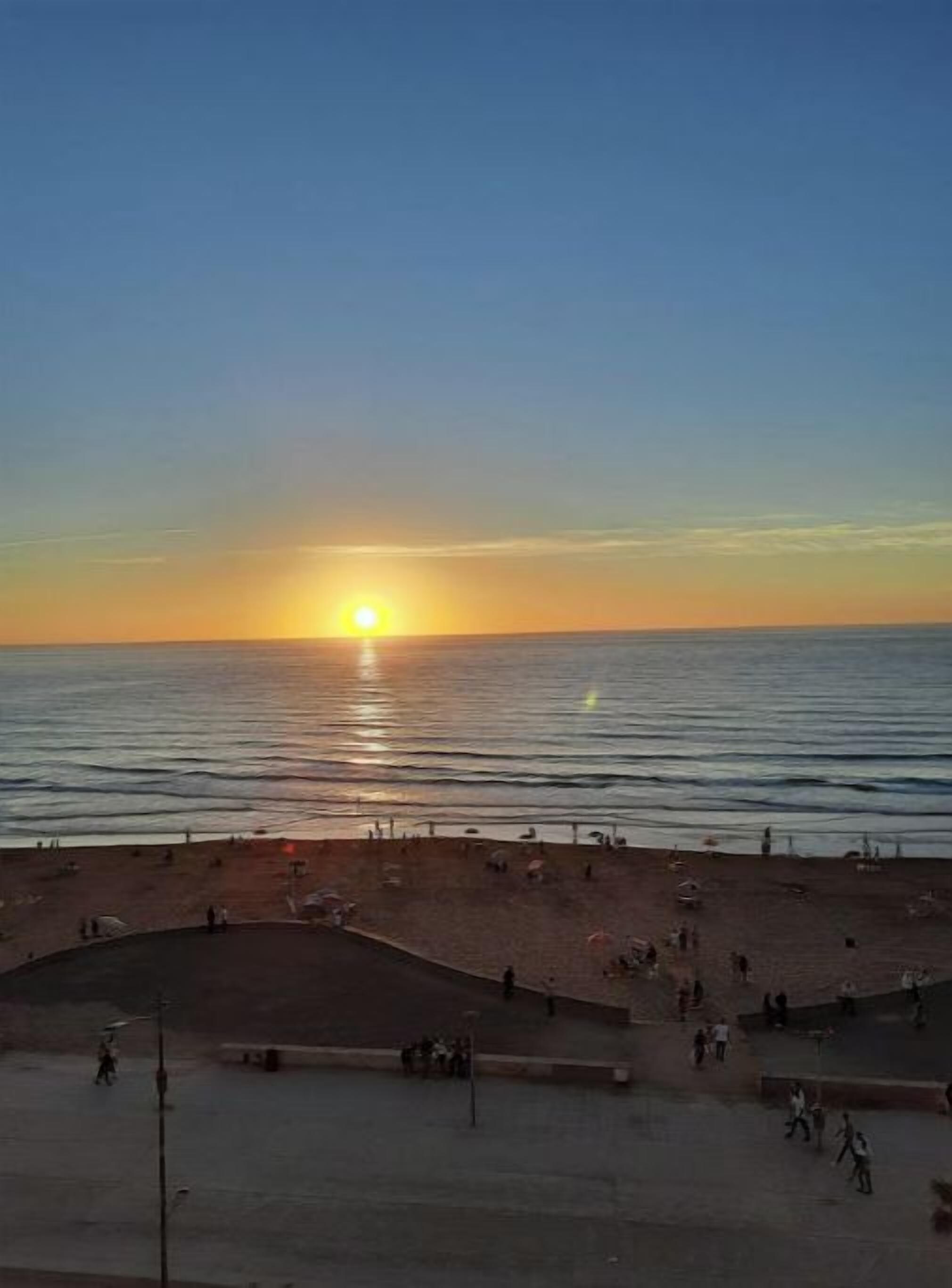 A beach with a sunset, people walking, and a paved area with benches.