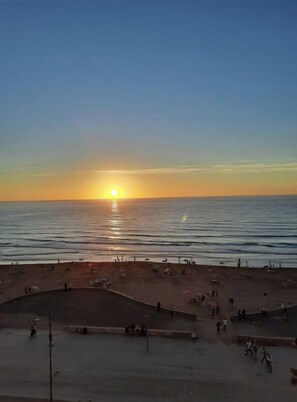 Une plage au coucher du soleil, avec des gens qui se promènent et une zone pavée dotée de bancs.