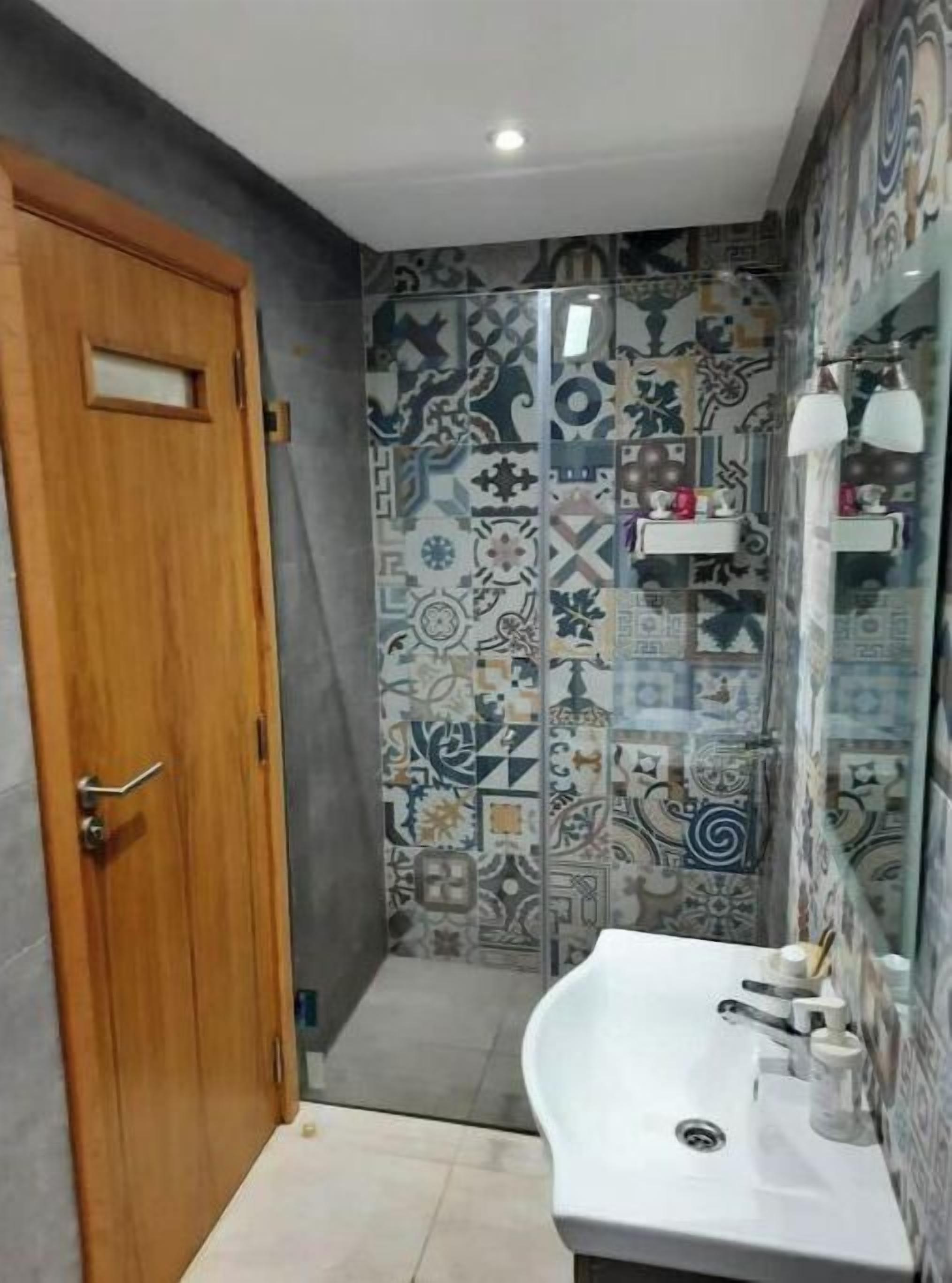 A bathroom with a wooden door, patterned tiles, a white sink, and a mirror with shelves.