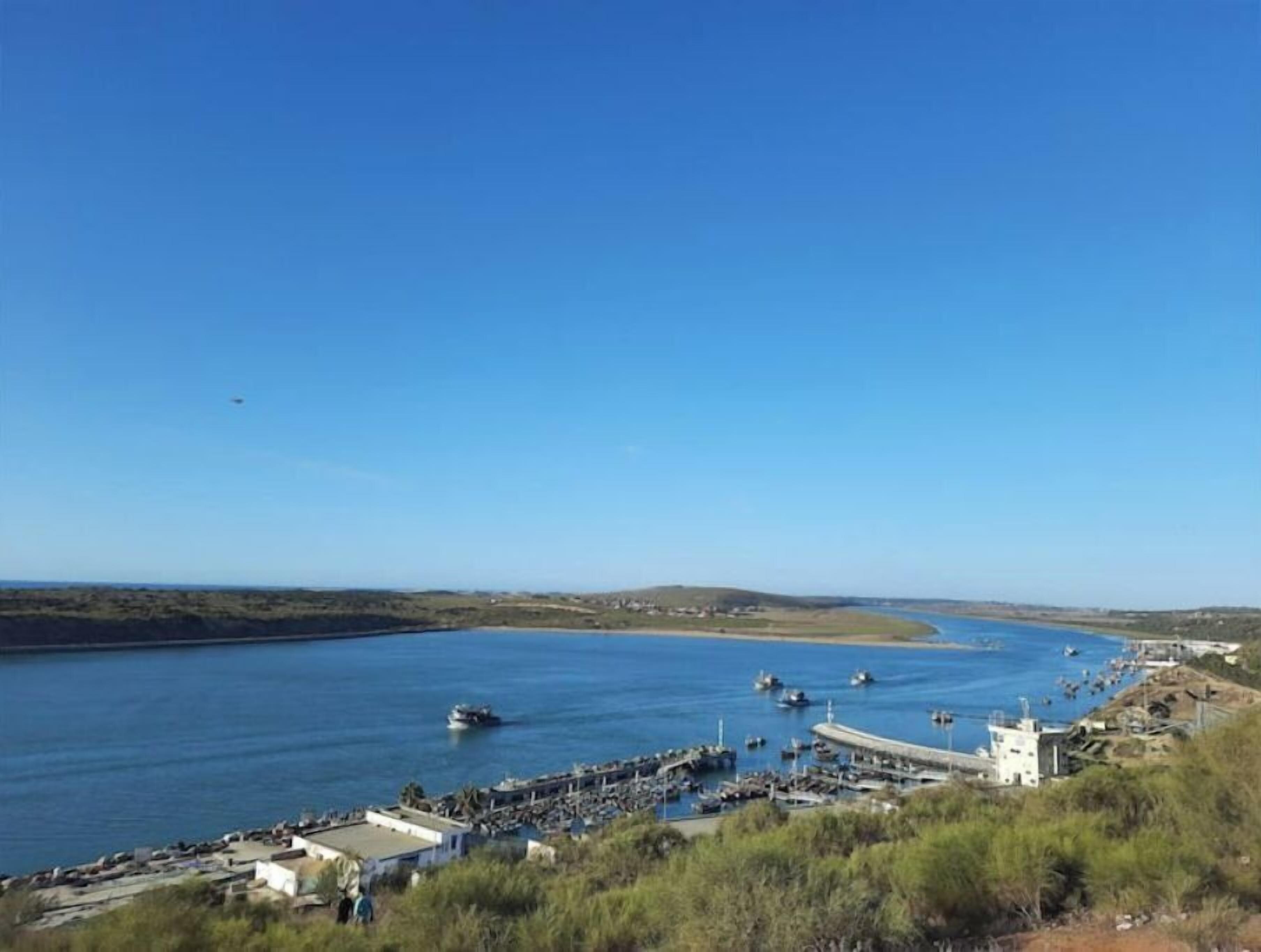Una marina costera con barcos amarrados, un muelle que se adentra en el agua y un cielo azul despejado.