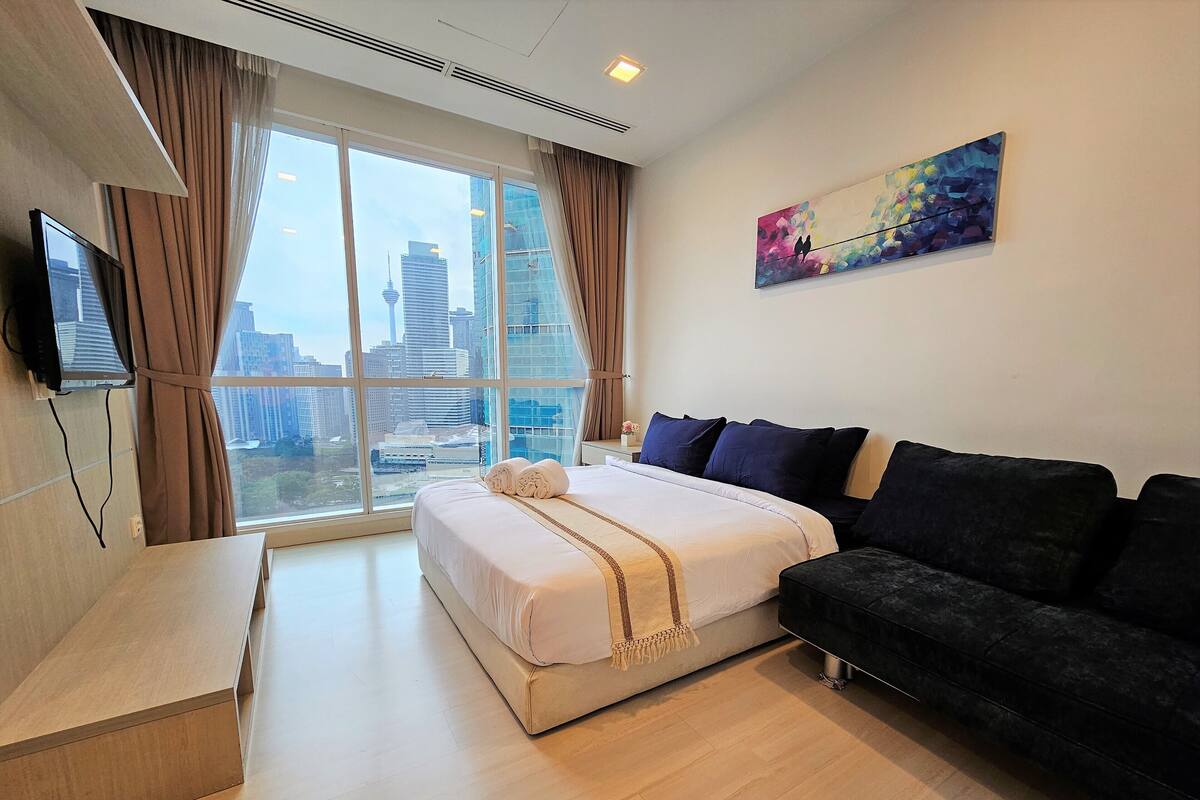 Habitación de hotel moderna con un ventanal que ofrece vistas a la ciudad, cama con ropa de cama oscura y clara, sof á negro y televisor de pantalla plana montado en la pared.