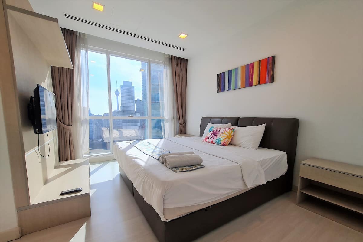 Habitación de hotel moderna con una cama grande, un televisor de pantalla plana montado en la pared y vistas a la ciudad a través de amplios ventanales.