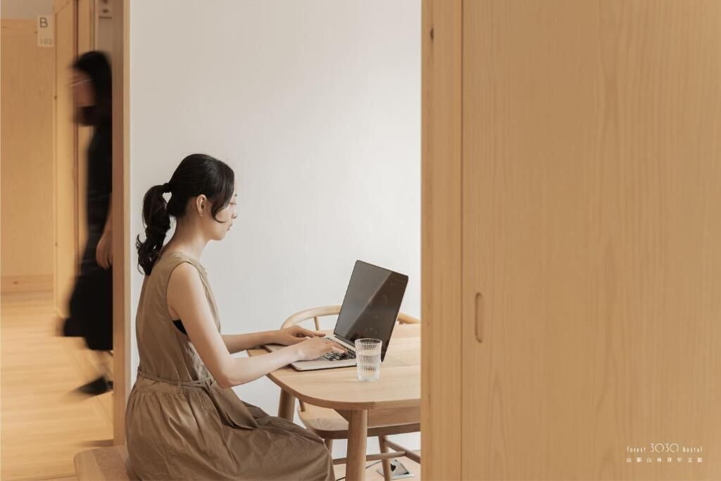 Une femme est assise à une table ronde en bois, devant un ordinateur portable, et un verre d’eau se trouve à côté d’elle.