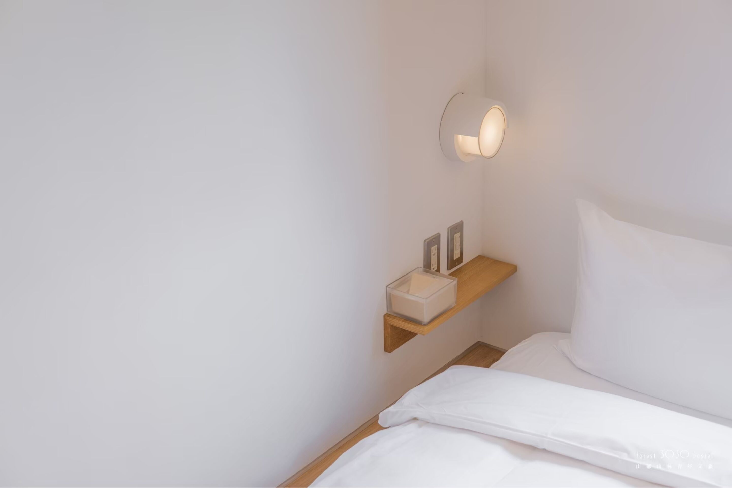 Cama bem arrumada com lençóis brancos, mesa de cabeceira com abajur e pequena estante com caixa e fotografias emolduradas.