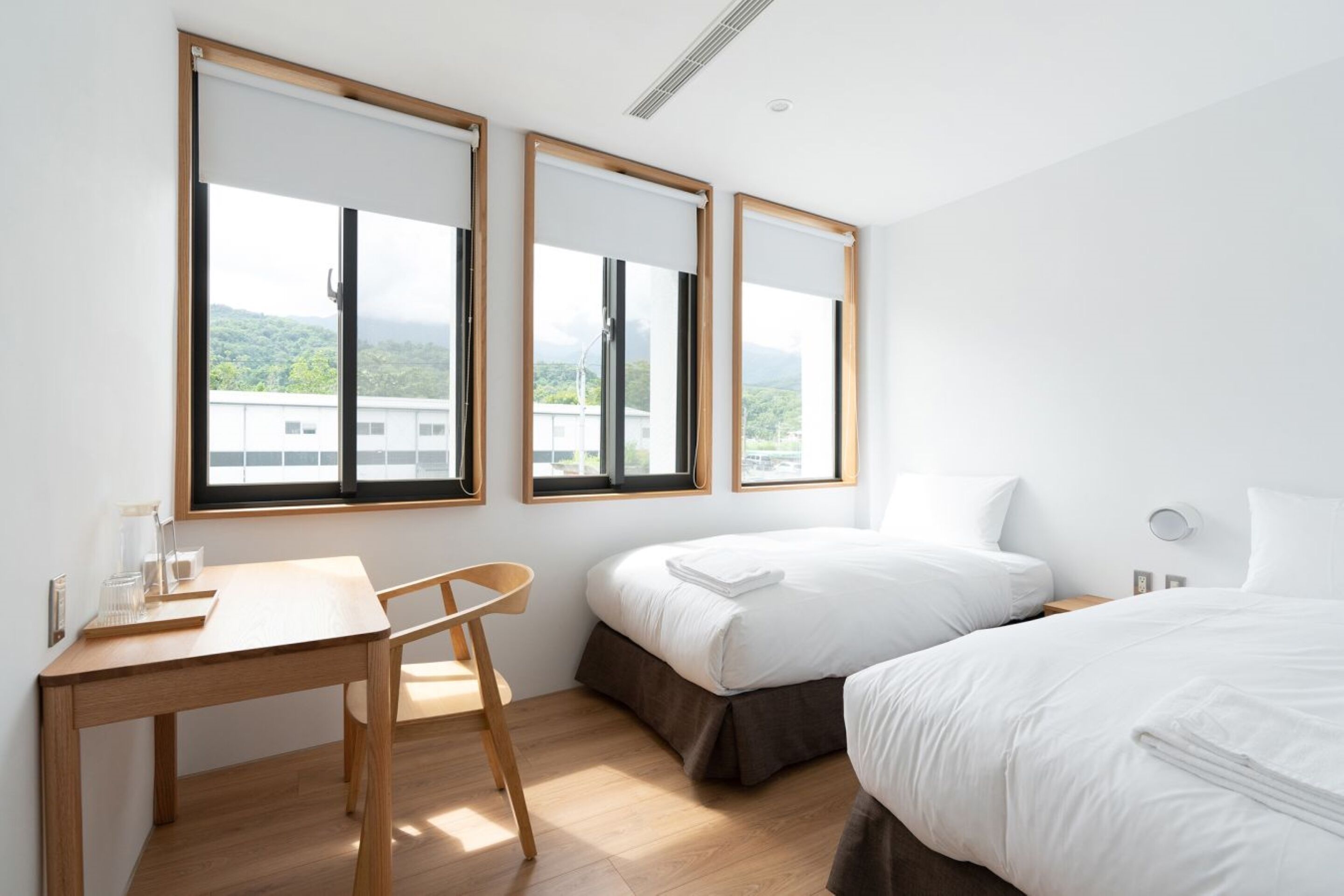 A hotel room with a wooden desk, two beds, and large windows with a view of buildings and greenery.