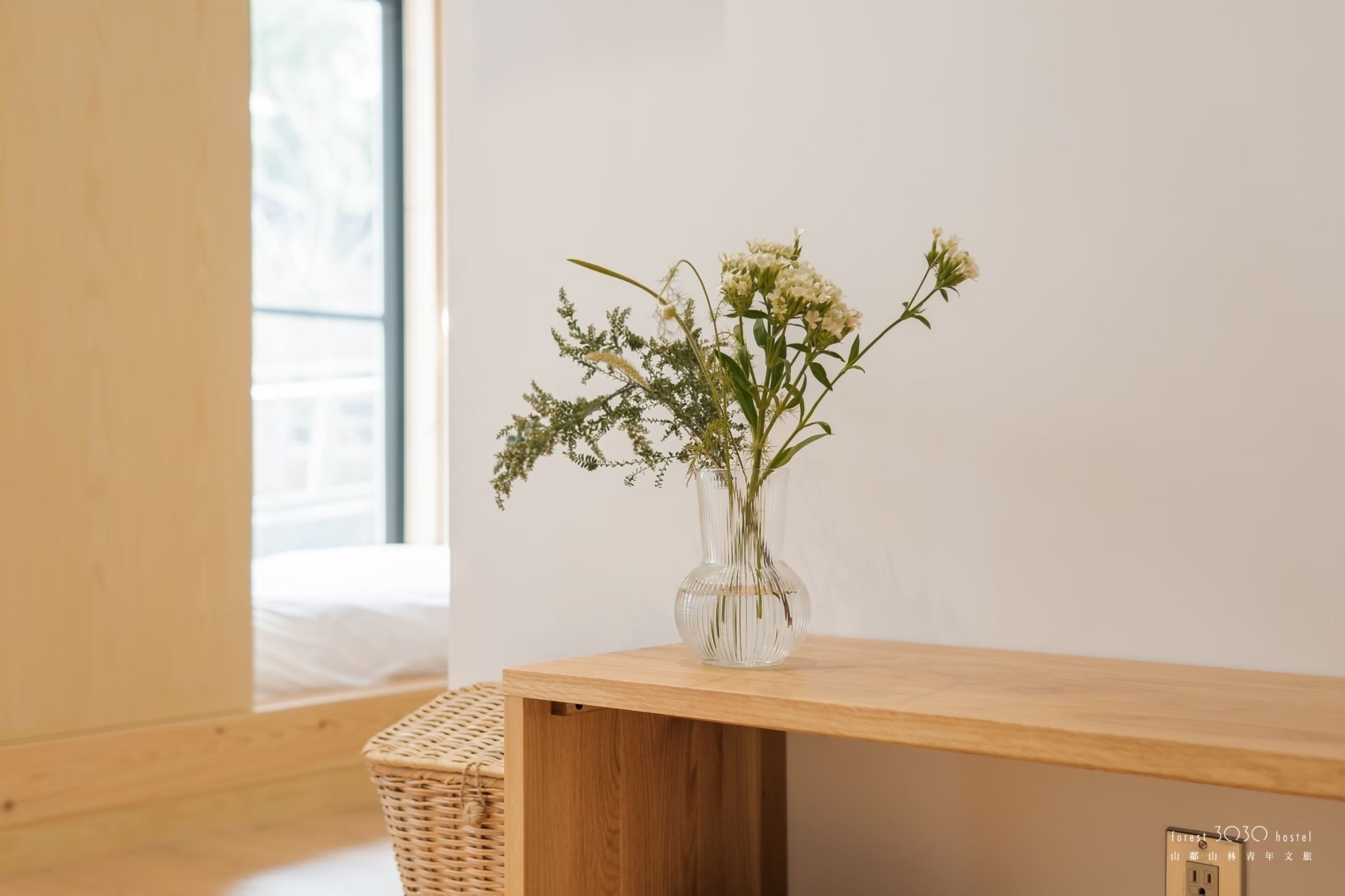 A wooden table with a clear vase containing white flowers, positioned against a plain wall with a window in the background.