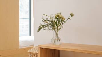 A wooden table with a clear vase containing white flowers, positioned against a plain wall with a window in the background.