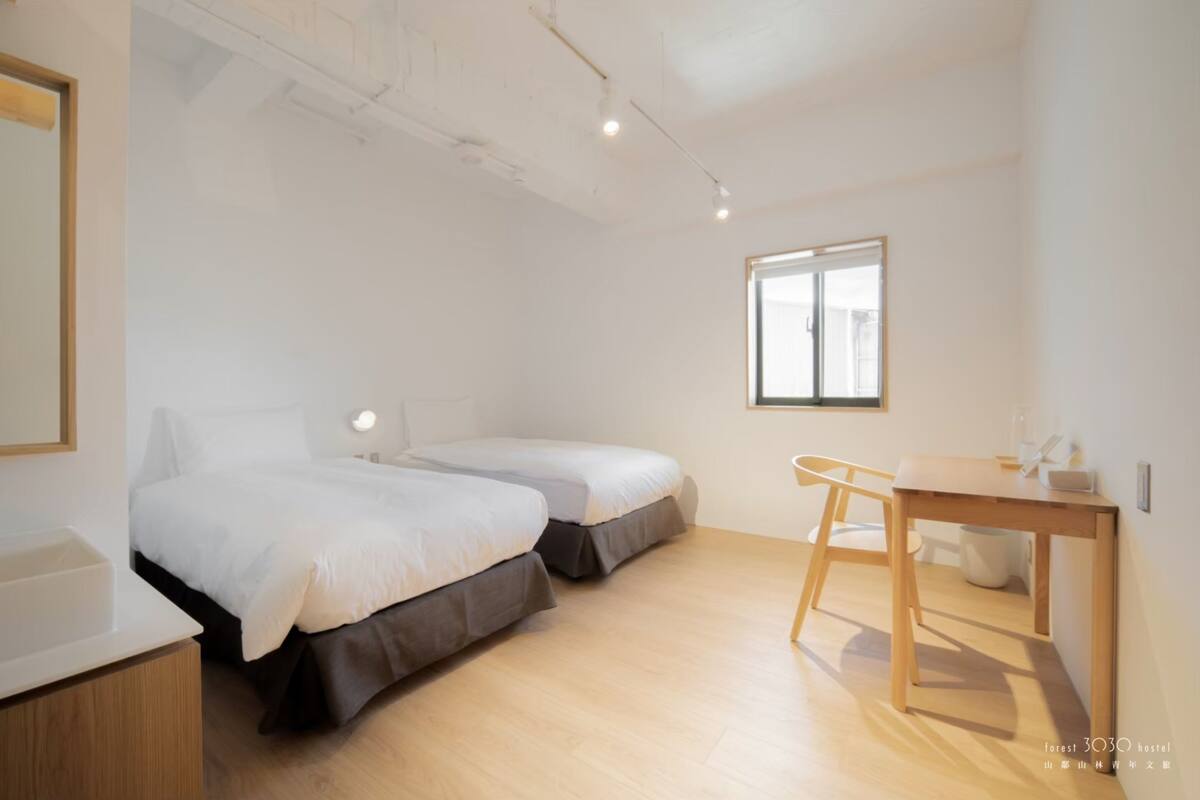 Una habitación limpia y moderna con dos camas, un escritorio de madera y un ventanal grande.
