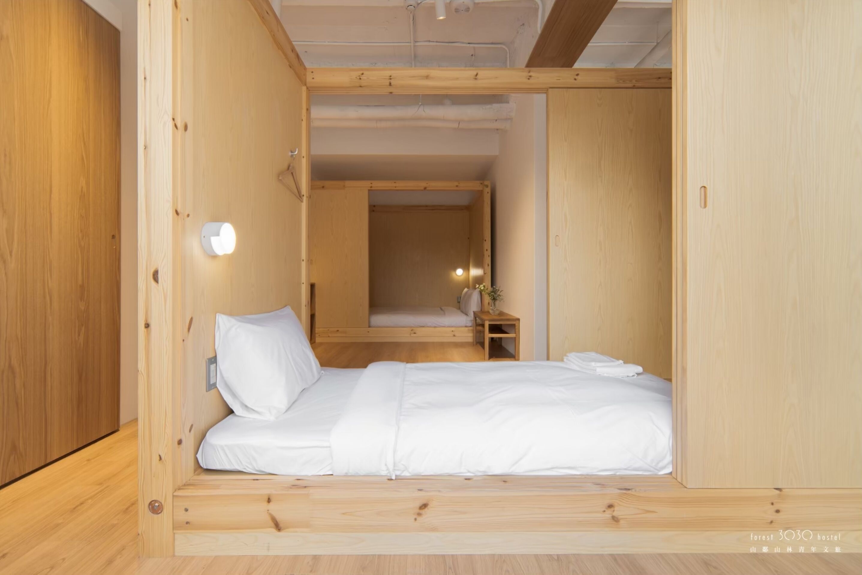 A compact wooden room with a bed, a nightstand, and a wall-mounted light.