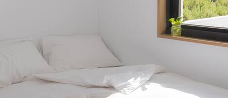 A neatly made bed with white linens, a book on the bed, and a small plant in a window ledge.