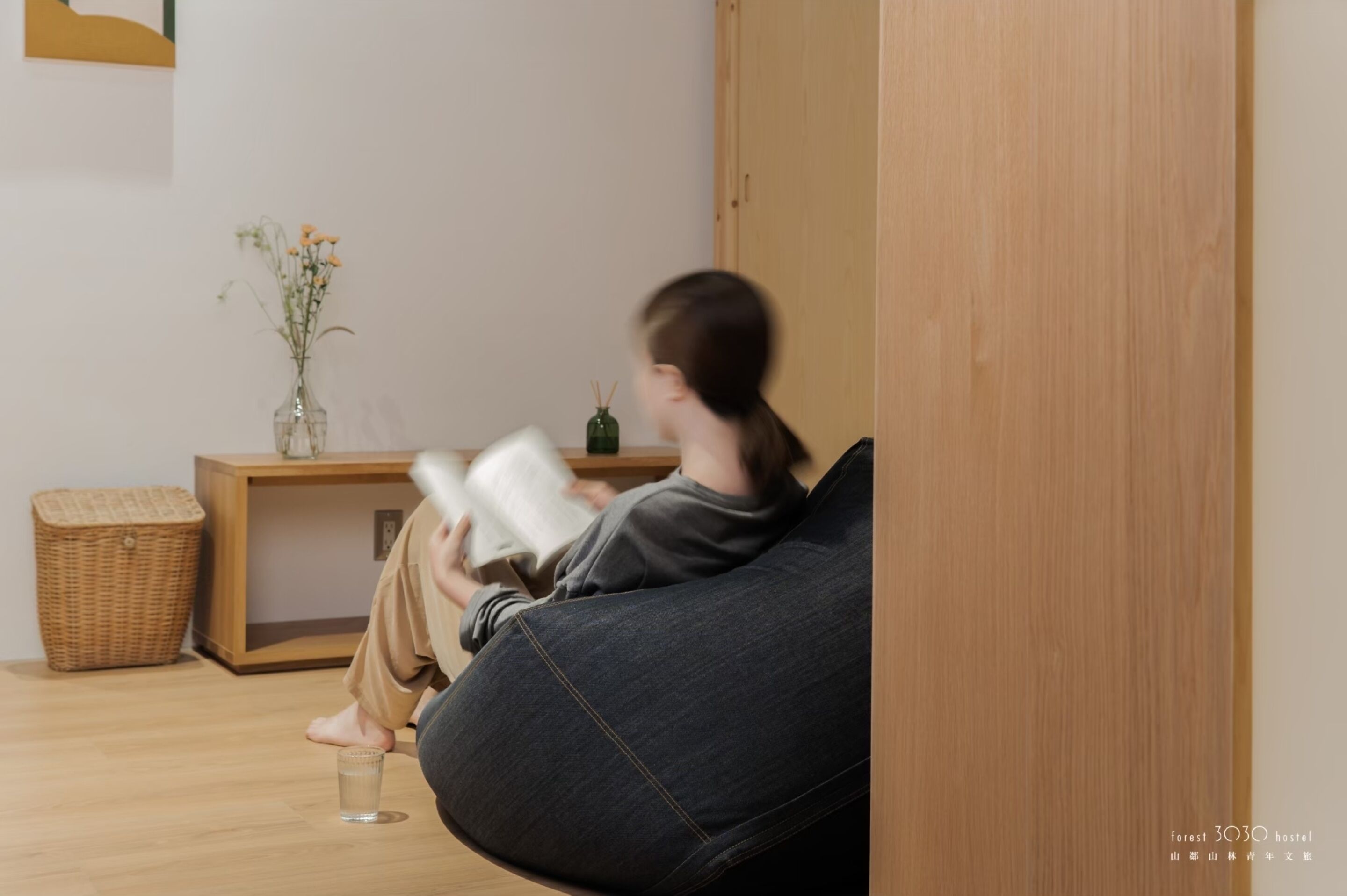 Une personne est assise sur un fauteuil en forme de sac de haricots, lisant un livre dans une pièce dont le sol est en bois, où se trouvent une petite table avec un vase de fleurs et un panier en osier.