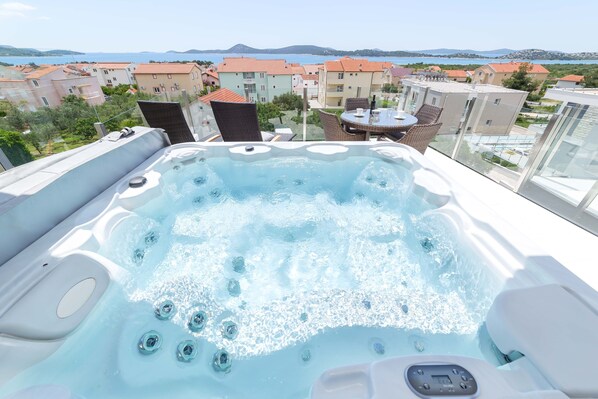 A hot tub with a control panel, surrounded by outdoor furniture, overlooking a coastal view with buildings and hills.