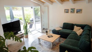 A living room with a green sofa, a coffee table, a TV, and a sliding glass door leading to an outdoor area with a patio umbrella. - Guest house Leonidas (Stromberg)