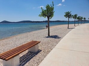 A pebble beach with a wooden bench and trees, a clear sky, and a distant mountain range. - Apartment with a Pool at Villa Aliara (Vodice)