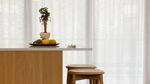 Eine Küche mit einer Holzinsel, zwei hölzernen Barhocker, einer hängenden orangefarbenen Pendelleuchte, einem Blumentopf und einem Fenster mit durchsichtigen Vorhängen.