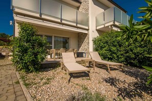 Un bâtiment moderne doté d’une façade en pierre, d’un balcon avec des garde-corps en verre et d’un espace extérieur aménagé avec des fauteuils et une table.