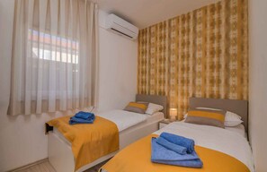 A hotel room with two beds, a window with sheer curtains, an air conditioning unit, and a patterned wall.