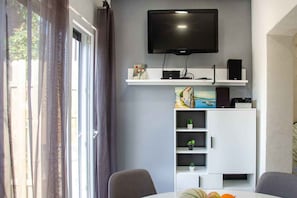 En moderne stue med et fladskærms-tv monteret på væggen, et hvidt tv-møbel med hylder og et spiseområde med bord og stole.