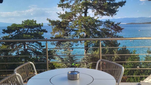 Ein runder Tisch mit Stühlen und einem Metallbehälter, mit Blick auf eine Küstenlandschaft mit Bäumen und Meerblick.