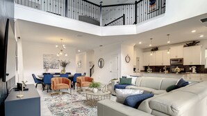 A spacious living area with a high ceiling, a dining area with blue chairs, a white sofa with blue cushions, and a kitchen with white cabinets. - Resort style villa close to Disney and other parks WALKING DISTANCE TO WATERPARK (Davenport)