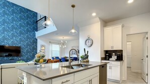 A modern kitchen with a central island, white cabinetry, and stainless steel appliances. There is a dining area with blue chairs and a flat-screen TV mounted on the wall. - Resort style villa close to Disney and other parks WALKING DISTANCE TO WATERPARK (Davenport)