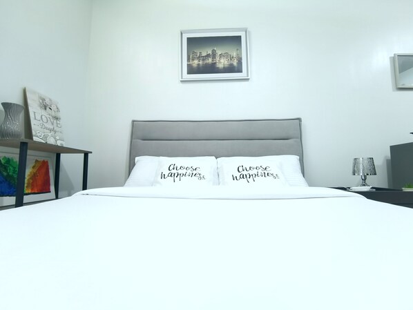 A neatly made bed with two pillows, a framed cityscape picture above it, and a small table with a vase and a framed artwork to the left. - COZY PLACE IN MACTAN W/ POOL, WIFI, NETFLIX - 10 min to the Airport! (Lapu-Lapu)