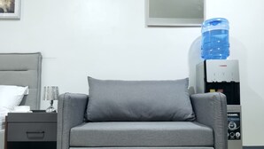 En grå sofa med en pude, en vanddispenser, en lampe og en seng med en hvid pude samt et natbord med en skuffe.