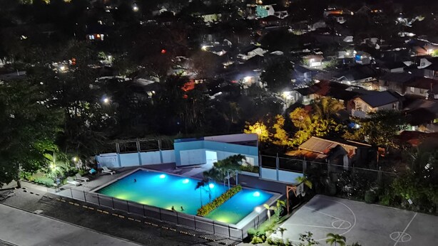 Oversigtsbillede af et boligområde med en swimmingpool omgivet af et hegn, træer og huse i baggrunden.