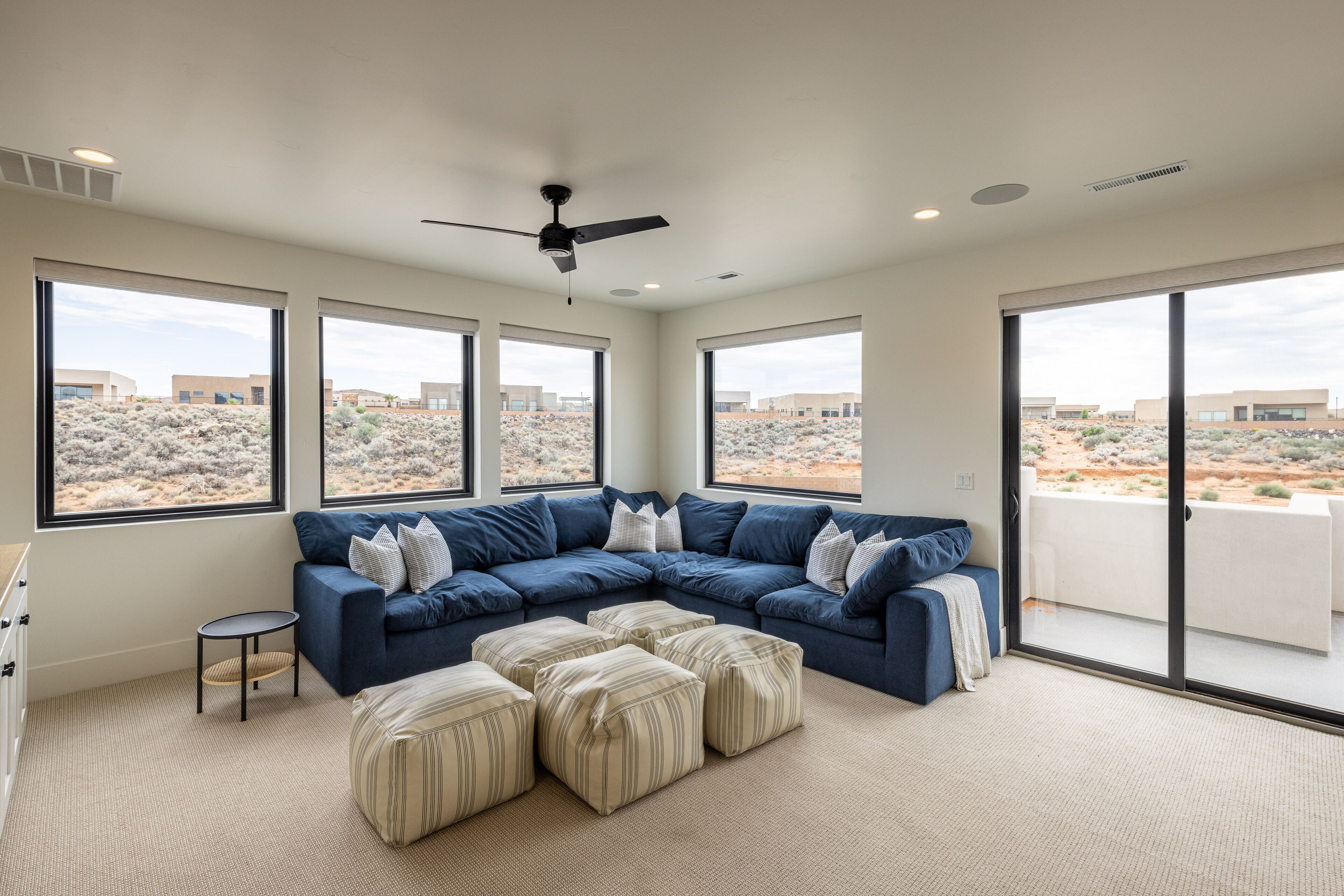 A modern living room with a sectional sofa, ottomans, a small side table, and large windows offering a view of a desert landscape.