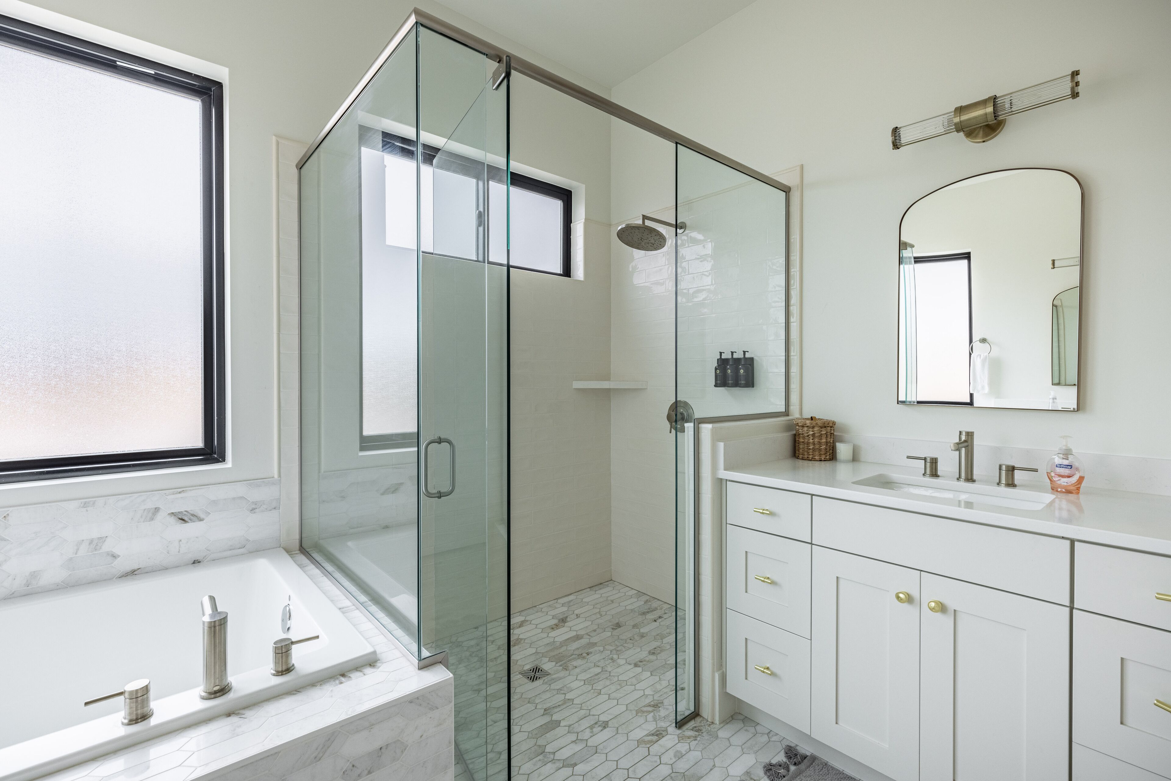 A modern bathroom with a glass-enclosed shower, a large window, a marble sink, and a white vanity with gold handles.