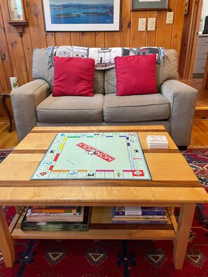 A living room with a grey sofa, two red cushions, a wooden coffee table with a Monopoly board game, and a bookshelf with various books.