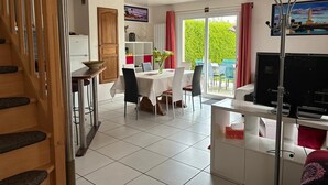 A room with a dining table, chairs, a TV, a staircase, and a view of greenery outside. - Audierne/Plouhinec: 1 km from the sea, in a peaceful, enclosed setting. (Plouhinec)