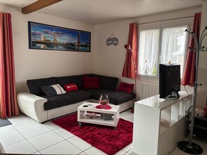 A living room with a black sofa, red cushions, a white coffee table, a painting of a coastal scene, and a TV on a stand.
