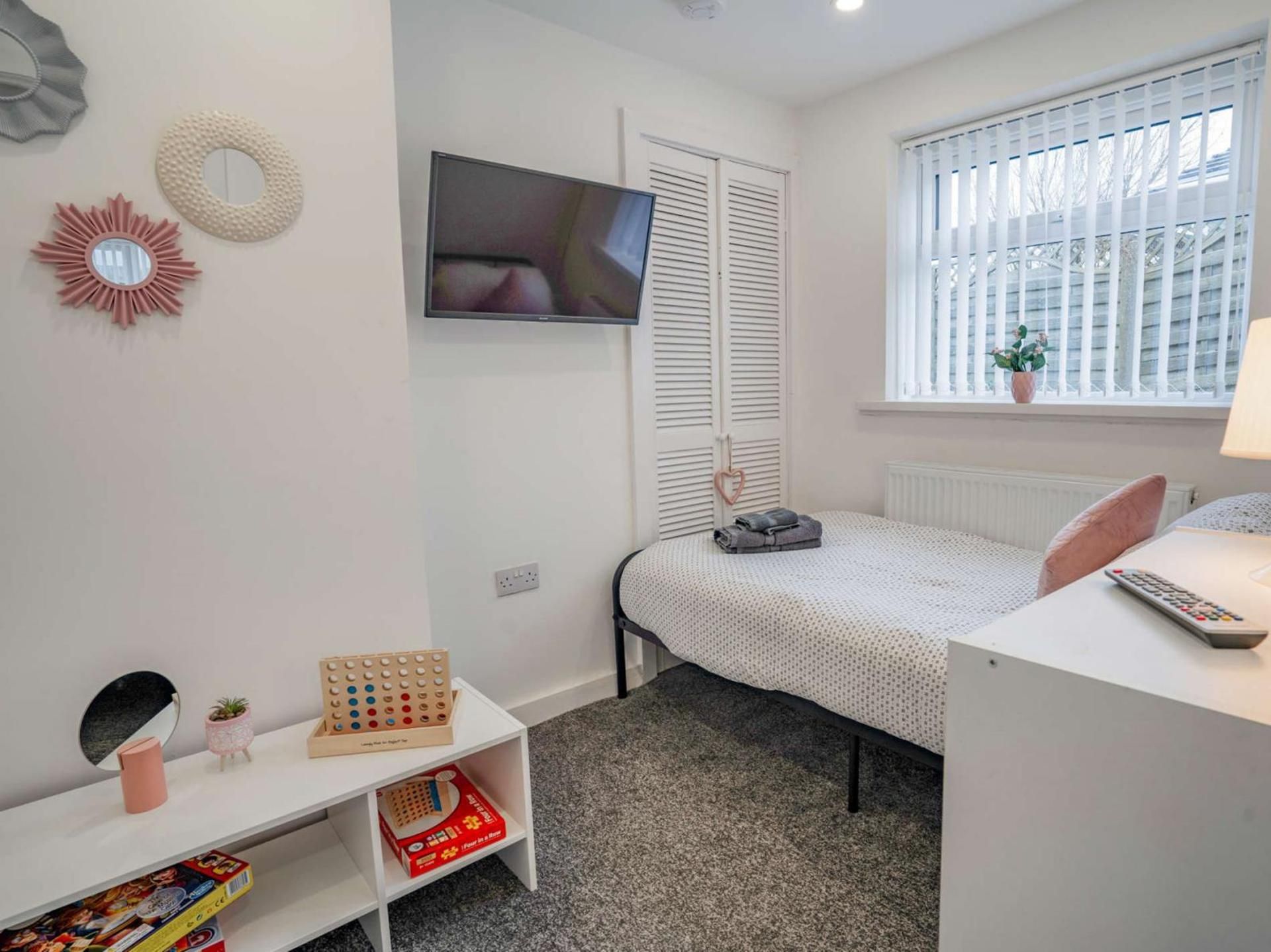 A compact bedroom with a bed, a TV mounted on the wall, a window with blinds, a small shelf with items, and a radiator.