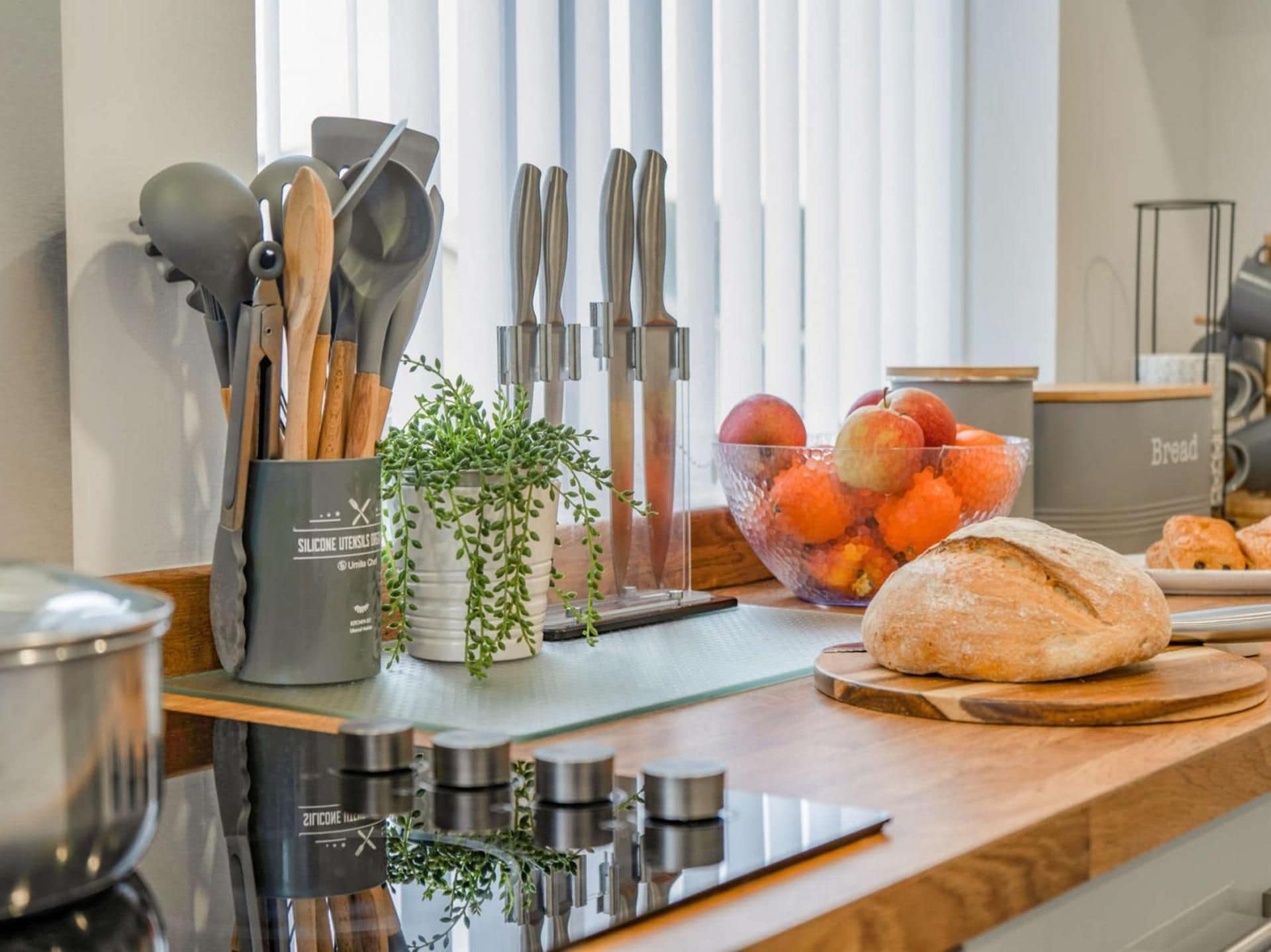 A kitchen counter with various utensils, a bowl of fruit, a loaf of bread, and a silicone utensil holder.