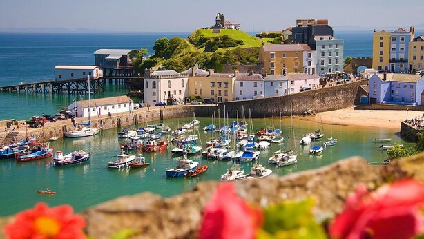 A coastal town with a marina filled with boats, historic buildings, and a prominent statue on a hill.
