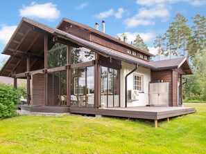 Ein modernes Haus mit großen Glasfenstern, einer Holzterrasse und einer kleinen Sitzgelegenheit im Freien.
