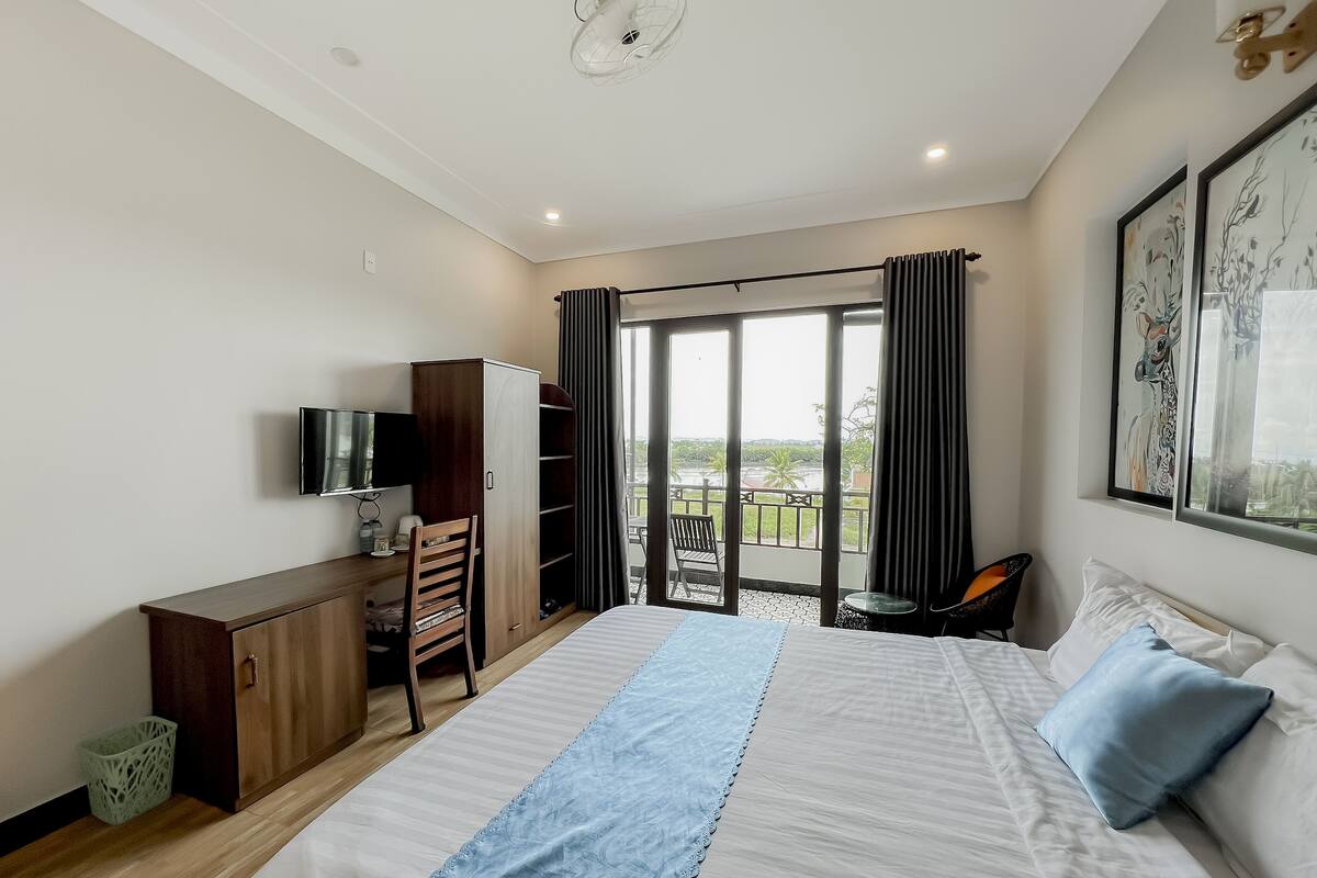 A hotel room with a large bed, a desk with a TV, a wardrobe, and a balcony with a view.