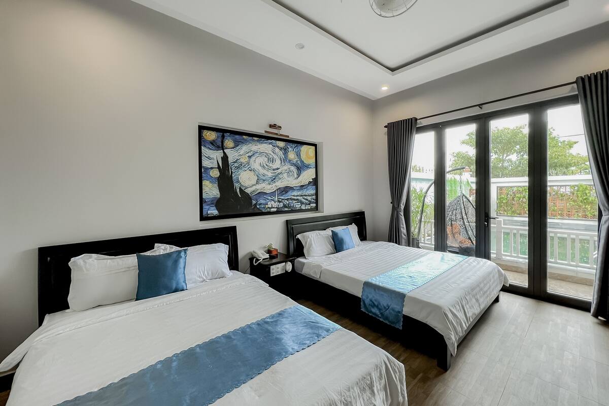 A hotel room with two beds, a ceiling fan, a large window with curtains, and a painting on the wall.