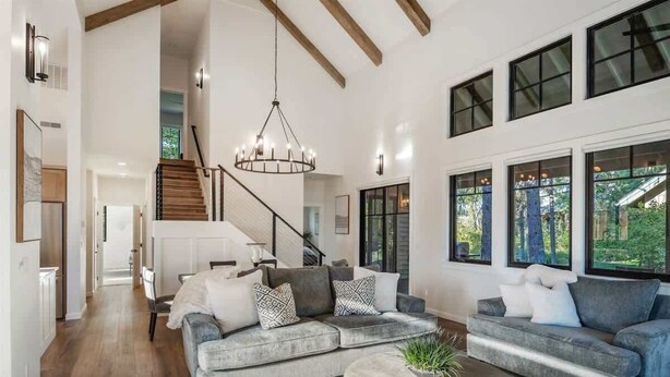 A spacious living room with a large gray sectional sofa, a wooden coffee table, and a chandelier. A staircase leads to an upper level, and large windows provide natural light and views of the outdoors. - Dover Bay Luxe Home w/ Private Hot Tub - Sleeps 12 (Dover)
